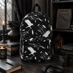 Peut inclure: Un sac à dos noir orné d'un motif répétitif blanc représentant des corbeaux en vol et des plumes éparpillées. Le sac comprend un compartiment principal et une poche frontale zippée, tous deux décorés du même motif monochrome d'oiseaux.