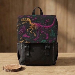 May include: Black backpack with a flap displaying a vibrant dinosaur skeleton design. The design incorporates orange, pink, and green elements against a black backdrop. The backpack features a black body, a top handle, and two front buckles.