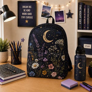 May include: A dark blue backpack featuring a celestial crescent moon and star pattern above a vibrant botanical print of lavender, daisies, and wildflowers. The backpack has a top handle, a front zippered pocket, and a main compartment with a zipper closure.