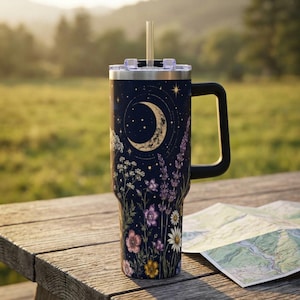 May include: A dark blue stainless steel tumbler with a handle and a clear straw. The exterior features a celestial design with a crescent moon, stars, and a variety of colorful wildflowers including lavender, daisies, and pink blossoms.