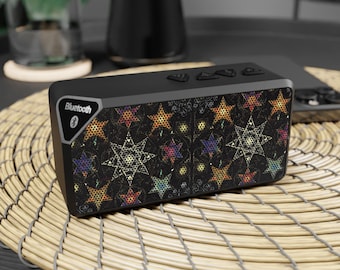 May include: Black rectangular Bluetooth speaker with a star pattern in multiple colors. The speaker has buttons on top and the Bluetooth logo on the side. It sits on a woven mat.