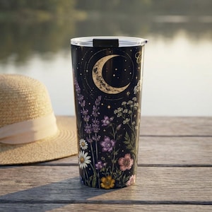 May include: A cylindrical tumbler with a black background featuring a gold crescent moon, stars, and a variety of colorful wildflowers including purple lavender, white daisies, and pink blossoms. The cup has a clear plastic lid with a black closure.
