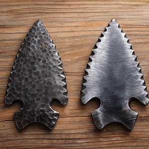 May include: Two metal arrowhead sculptures with a rustic finish rest on a wooden surface. One arrowhead has a textured, hammered appearance, while the other has a smooth, brushed metal look. The arrowheads are dark gray.