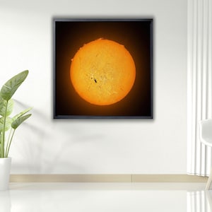 Può includere: Un'immagine incorniciata del sole, una sfera giallo-arancio brillante con macchie scure, su uno sfondo nero. L'opera d'arte è esposta su una parete bianca in un ambiente interno moderno, con piante e una sedia bianca.