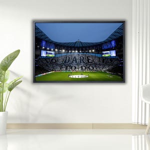 Könnte beinhalten: Gerahmtes Stadionbild mit grünem Feld und einer Menge, die die Worte "DARE TO DO" zeigt. Das Stadion hat helle Lichter und einen dunkelblauen Himmel. Das Bild hängt an einer weißen Wand.