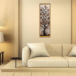 May include: A framed wall art piece featuring a stylized tree with white, brown, and copper-colored leaves. The artwork is set against a white background and framed in gold, displayed above a beige sofa in a modern living room.