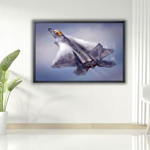 Op de afbeelding: Ingelijste print van een grijze en zilveren F-22 Raptor straaljager in vlucht, met condenssporen tegen een blauwe lucht. Het vliegtuig is omhoog gekanteld en toont zijn kenmerkende ontwerp. Het kunstwerk wordt aan een witte muur getoond.