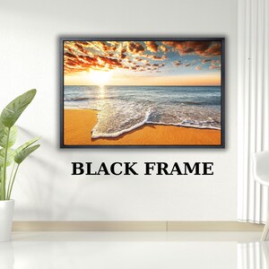 Puede incluir: Una pieza de arte mural rectangular que muestra una puesta de sol sobre el océano con arena dorada y olas, encerrada en un marco negro. El texto BLACK FRAME está impreso en negro debajo de la obra, que representa un sol brillante, nubes naranjas y agua azul.