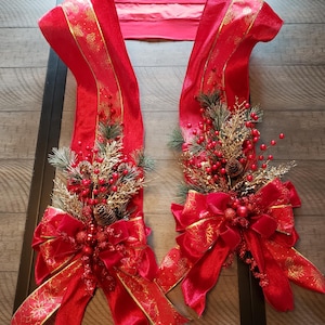 May include: A festive red Christmas swag with gold accents. The swag features decorative bows, pinecones, berries, and greenery. The design is symmetrical, with a central connecting piece.
