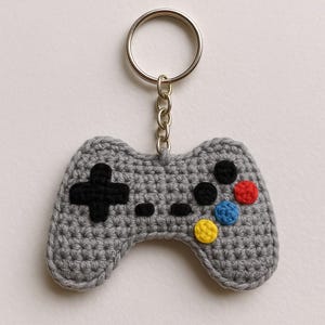 Peut inclure: Un porte-clés en forme de manette de jeu vidéo grise crochetée, avec un anneau argenté. La manette présente une croix directionnelle noire, des boutons noirs et des boutons colorés en rouge, bleu et jaune.