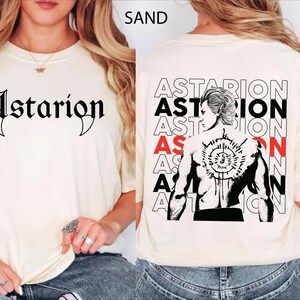 May include: A sand-colored t-shirt featuring the text Astarion in black gothic lettering on the front. The back displays a graphic of a character with a detailed circular tattoo on their back, surrounded by the repeated text Astarion in black and red.