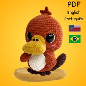 May include: A handmade crochet platypus toy in shades of brown, yellow, and white, with large black eyes. The toy has a yellow beak, a white belly, and a brown tail. The image includes the text "PDF English Português" and flags of the USA and Brazil.