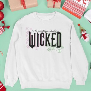 Puede incluir: Sudadera blanca con la frase "The weather outside is WICKED" en letras negras y rosas. La imagen está rodeada de decoraciones navideñas, como adornos, regalos y un gorro de Papá Noel.