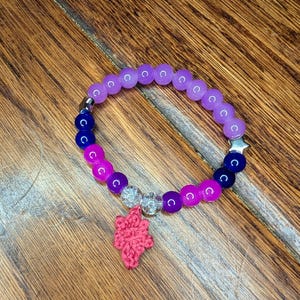 Op de afbeelding: Een armband met paarse, blauwe en roze kralen, een zilveren sterbedel en een roze gehaakte bladbedel. De armband ligt op een houten oppervlak. De kralen hebben een diameter van ongeveer 0,8 cm.