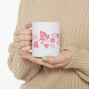 May include: White ceramic mug featuring a pink floral design with leaves, berries, and star-shaped flowers. The mug is held by a person wearing a beige sweater. The design wraps around the mug.