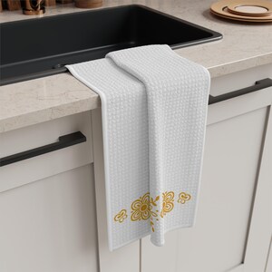Può includere: Asciugamano da cucina bianco con trama strutturata e motivo floreale dorato. L'asciugamano è piegato su un piano di lavoro, mostrando il suo design e la combinazione di colori. Realizzato in materiale morbido e assorbente.