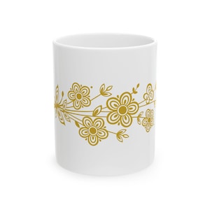 May include: White ceramic mug with a gold floral pattern. The design shows stylized flowers and leaves arranged horizontally around the mug. The mug is cylindrical and has a smooth, glossy finish.