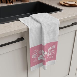 Può includere: Asciugamano da cucina bianco con una fascia rosa con un motivo floreale bianco. L'asciugamano è piegato su un piano di lavoro, mostrando la sua trama strutturata e il suo motivo decorativo. L'asciugamano è realizzato in un materiale morbido.