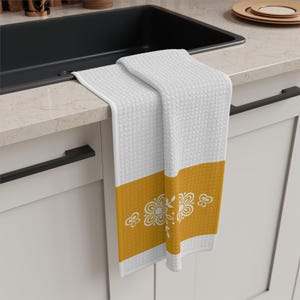 Può includere: Asciugamano da cucina bianco con trama strutturata e una fascia giallo senape con disegni floreali bianchi. L'asciugamano è piegato su un piano di lavoro, mostrando il suo motivo decorativo e la combinazione di colori.