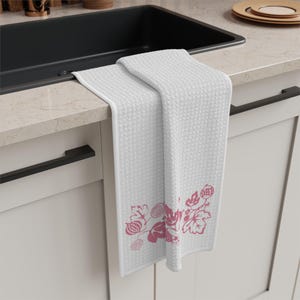 Può includere: Asciugamano da cucina bianco con trama a rilievo e un disegno floreale rosa nella parte inferiore. L'asciugamano è piegato su un piano di lavoro, mostrando la sua estetica pulita e semplice. L'asciugamano è appeso sopra un lavandino nero.