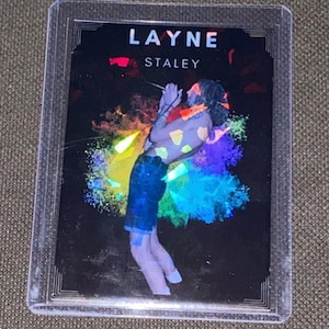 May include: A trading card featuring Layne Staley, with the name in white at the top. The image shows a colorful, artistic depiction of Staley singing into a microphone, with a rainbow-colored burst behind him. The card is encased in a protective sleeve.