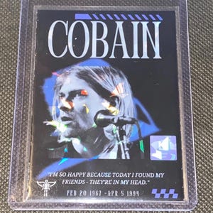 May include: A collectible card featuring the name "COBAIN" in white capital letters above a portrait of a musician. The card has a black background with blue and yellow accents. Text includes a quote and dates.