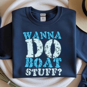 Peut inclure: Sweat-shirt bleu marine avec l'inscription "WANNA DO BOAT STUFF?" en bleu clair et blanc avec un effet vieilli. Le sweat-shirt est présenté sur une surface blanche, avec un chapeau de paille en arrière-plan.