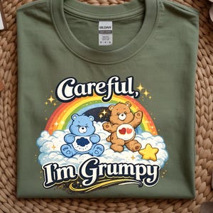 Könnte beinhalten: Olivgrünes T-Shirt mit einer Cartoon-Grafik von zwei Bären, einem Regenbogen und dem Text "Careful, I'm Grumpy". Die Bären sind braun und blau, mit einem gelben Stern. Das Shirt ist aus weichem Material.