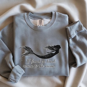 Puede incluir: Sudadera azul claro con una silueta de sirena negra. La sirena tiene pelo largo y ondeante y una cola detallada. El texto "If found on land, return to the ocean" está impreso debajo.