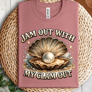 May include: A mauve t-shirt featuring a graphic of an open clam with a large white pearl inside, surrounded by water splashes and sparkles. The text "JAM OUT WITH MY CLAM OUT" is printed in a bold, arched font above and below the illustration.
