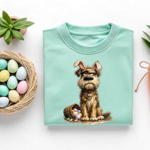 May include: A light teal sweatshirt featuring a grumpy dog wearing bunny ears and a neckerchief. The dog sits beside a basket of colorful Easter eggs. A fun, whimsical design for the spring season.