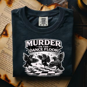 May include: Dark navy blue t-shirt with the text "MURDER ON THE DANCE FLOOR" above a graphic of three dancing crows on a black and white checkered floor. The shirt has a crew neck and a tag that says "COMFORT COLORS".