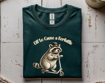 Può includere: T-shirt verde scuro con un'illustrazione di un procione su uno scooter, con un cappello a bombetta. Il testo "Off To Cause a Kerfuffle" è arcuato sopra. La maglietta è piegata su una superficie di legno chiaro.