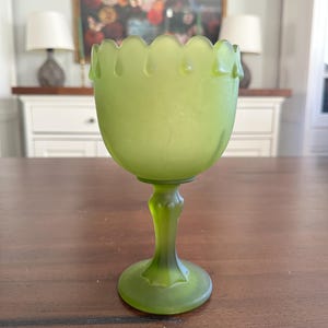May include: A green glass goblet with a scalloped rim and a detailed stem. The goblet has a rounded bowl shape and sits on a circular base. The glass has a matte finish and a light green color.
