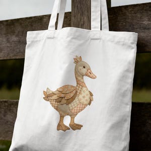 Puede incluir: Un bolso tote blanco con una aplicación decorativa de un pato. El pato tiene un cuerpo beige con detalles de patchwork en varios patrones y colores, incluyendo marrón, beige y rosa. El bolso está colgado de una valla de madera.