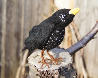 May include: A black felt bird with a yellow beak and brown legs. The bird is perched on a tree stump.