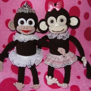 Puede incluir: Dos muñecos de mono hechos a mano con ganchillo. Uno lleva una tiara plateada y un tutú de encaje blanco, el otro un lazo rosa y un tutú de encaje rosa. Ambos tienen cuerpos marrones, manos y pies color canela y ojos grandes. Posan sobre un fondo rosa de lunares.