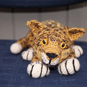 Puede incluir: Un animal de peluche de crochet que se asemeja a un leopardo, acostado sobre una superficie de tela azul. El animal tiene manchas negras y marrones, patas blancas y ojos amarillos.