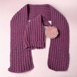 May include: A matching knitted scarf and hat set in a deep purple hue. The scarf is long and rectangular, while the hat has a pointed top and a fluffy pink pom-pom. The set is made of a thick, textured yarn.