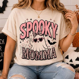 May include: A beige short-sleeve t-shirt featuring a graphic print with the text SPOOKY MOMMA in pink and black. The design includes a ghost wearing heart-shaped sunglasses and a leopard print bow, surrounded by stars, a spiderweb, and a bat.