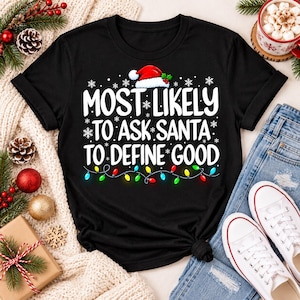 May include: A black short-sleeved t-shirt featuring a graphic with a Santa hat, snowflakes, and a string of colorful Christmas lights. The text reads: MOST LIKELY TO ASK SANTA TO DEFINE GOOD. The design is centered on the front of the garment.