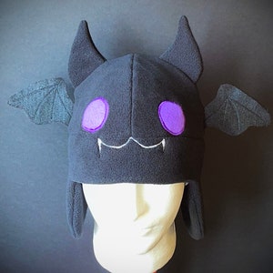May include: A black fleece hat shaped like a bat, with purple eyes and a white stitched mouth. The hat has bat ears and wings, and is displayed on a mannequin head. The hat is a costume accessory.