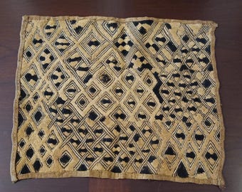May include: A rectangular textile with a repeating geometric pattern in black and tan. The design features diamonds, squares, and intersecting lines. The edges of the textile are finished with a simple border.