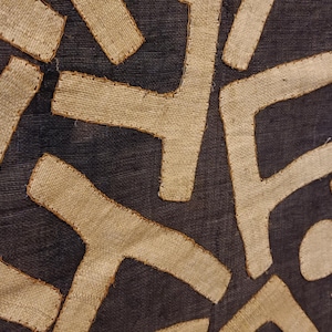 May include: Close-up of a dark blue textile with a repeating pattern of beige, angular shapes. The shapes are stitched onto the fabric, creating a textured surface. The design is geometric and abstract.