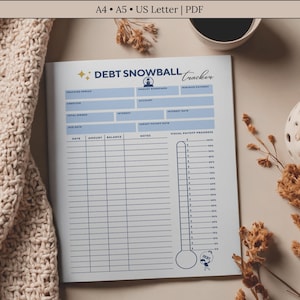 Op de afbeelding: Een afdrukbare "Debt Snowball Tracker" op een witte pagina met blauwe accenten. De pagina is open op een beige oppervlak, met een beige gebreide deken aan de linkerkant. De tekst "Debt Snowball Tracker" staat bovenaan. Een kopje koffie en decoratieve items zijn ook zichtbaar.