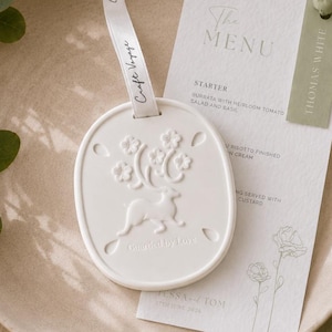 Puede incluir: Un adorno ovalado de cerámica blanca con un relieve de ciervo y flores, suspendido por una cinta blanca con la etiqueta Craft Voyage. Junto a él hay una tarjeta de menú blanca con detalles en verde salvia, con el texto The Menu y Thomas White.