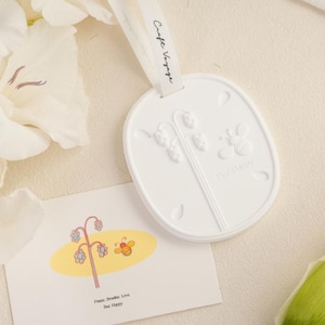 May include: A white oval ceramic hanging ornament featuring an embossed lily of the valley and bee design with the text Bee Happy. A white ribbon is attached to the top. A rectangular card displays a colorful illustration of a flower and bee.