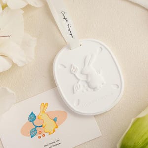 Könnte beinhalten: Ein weißes, ovales Keramikornament mit einem geprägten Hasen- und Blattmotiv, aufgehängt an einem weißen Band mit dem Aufdruck Craft Voyage. Eine rechteckige Karte zeigt eine gelbe Hasenillustration und den Text Pause, Breathe, Love, Hop into Happiness.