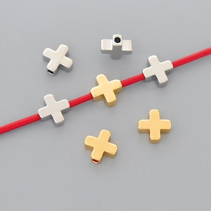 May include: A set of gold and silver plus sign shaped beads. The beads are strung on a red cord.