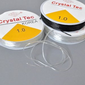 May include: Spools of clear and black elastic string, labeled "Crystal Tec KOREA 1.0". The clear string is unwound, showing its flexibility. The spools have a white label with red text and a yellow triangle.